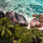 Aerial drone view of a woman standing on a tropical beach with palm tress and rock formations at Anse Source d'Argent, La Digue, Seychelles